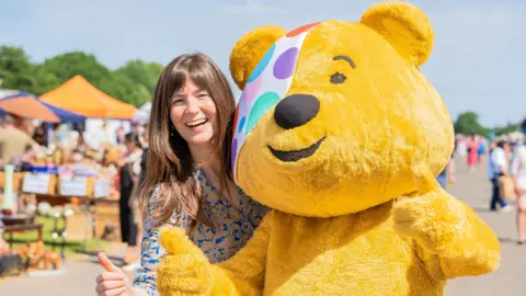Natasha and Pudsey in a close-up shot smiling to camera with thumbs up. The antiques fair is visible in the background with people milling about in the sunshine