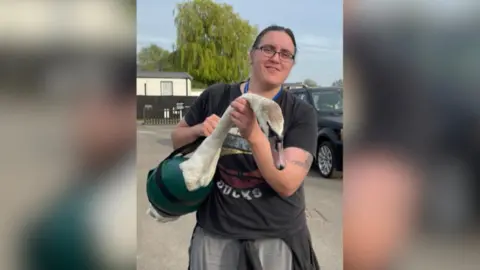 Abi Edis Abi Edis looks at the camera she is holding a swan in a sling. She has brown hair tied back and wears glasses. She is wearing a black t-shirt and grey jogging bottoms. 