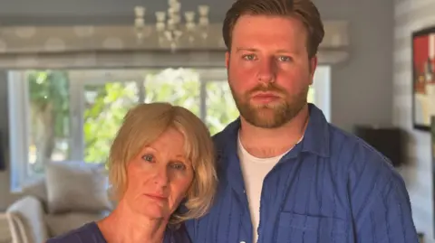 Paul's wife Carys stands on the left with blonde hair, her son is on the right, taller and wearing a blue shirt. Both are staring directly into the camera.
