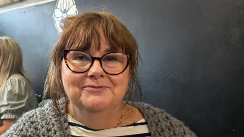 Ben Schofield/BBC Jane Maxwell, sitting in a coffee shop, with glasses on, with long hair tied back wearing a blue striped top and a cardigan. 