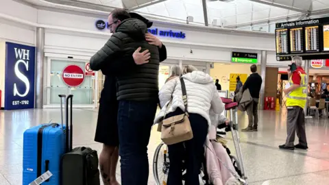 PA Media People with suitcases are hugged by loved ones in Stansted Airport's arrivals lounge, with the arrivals board visible behind them.