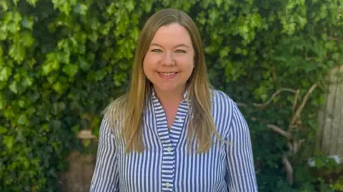 BBC A woman with blonde hair in a blue and white striped shirt stands smiling in front of green leafy bushes.