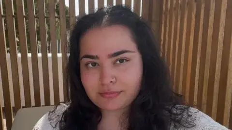 Romz Romz is taking a selfie-style photograph, looking directly at the camera. She has dark hair, brown eyes and a piercing in her nose. She is in front of a brown background and is wearing a white jumper.