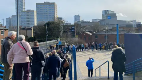 A queue of people stand in the sunshine as they wait to get into the indoor arena. City tower blocks can be seen in the background