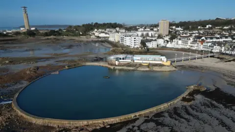 The lido at Havre des Pas at low tide on a clear blue sky day. The lido is circular with blue water and it is empty. Behind it are buildings and a high rise. To the left is a lighthouse.