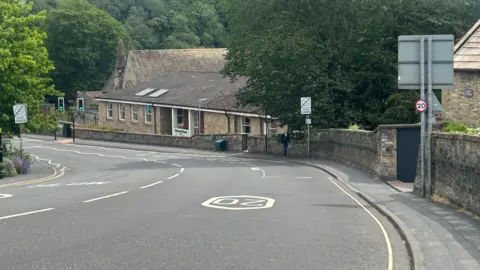 Mercury House, a large building on the side of a hilly road with lots of trees in the background and a 20 miles-per-hour speed restriction sign painted on the road.