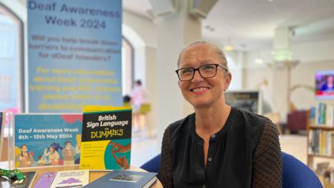 Carol smiles at the camera as she sits on a chair and on a table to the left of her is a table with books on British Sign Language and promotional art of Deaf Awareness Week