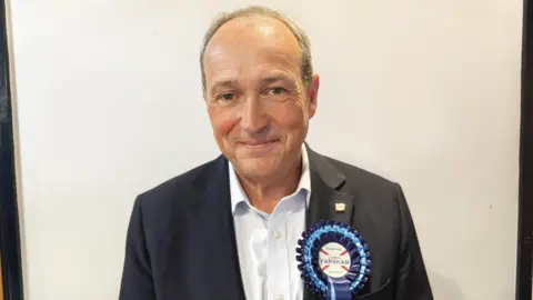 Lyndon Farnham smiles while standing in front of a white wall. He is wearing a black blazer and a white shirt. He has a Jersey flag badge on his left lapel. A large dark and sky blue rosette is on the same lapel.