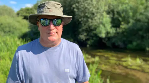 Owen Sennitt/BBC A man wearing a brimmed hat, sunglasses and a T-shirt looks towards the camera, with a stretch of river visible in the background on a warm summer's day