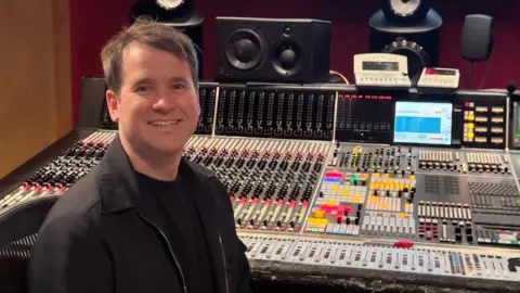Chris Peabody with short brown hair, a black top and black coat, seated at a large professional audio mixing console in a recording studio. The console is covered with rows of faders, knobs, buttons, and illuminated controls. Multiple studio monitor speakers are positioned around the workstation, and two small display screens sit above the mixing desk. The room features dark red walls and warm lighting, highlighting the extensive audio production equipment.