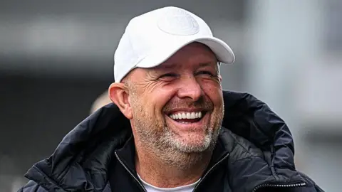 Andy Woodman looks to his left and smiles. He is wearing a white cap and black coat with hood