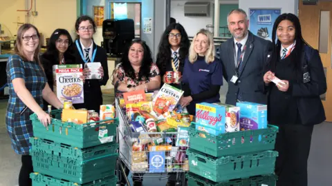Lawn Manor Academy A group of four adults and four children in school uniform pose behind a trolley and green crates stuffed with food items including Cheerios cereal. They are all smiling.