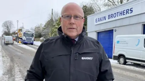 Shows Peter Eakin wearing a company coat, standing on the roadside with traffic in the background. He is balding with black-framed glasses.