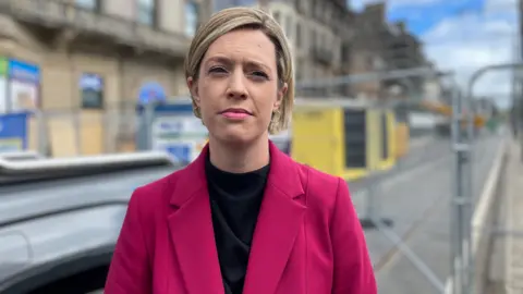Jenny Gilruth looking straight at the camera. She is wearing a pink jacket over a black top. She has short blonde hair. She is standing in front of a blurred Princes Street with a heras fence and a building site behind her.