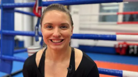 Kat Samuelson pictured at the Rittijak Gym in Swanscombe. Kat is wearing a black top and is smiling to camera. She is sat outside a boxing ring at the gym.
