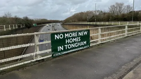 A sign is posted to a fence on a bridge going over the A331, which says 'NO MORE NEW HOMES IN TONGHAM' in capital letters. Cars travelling along the A331 can be seen in the background. 