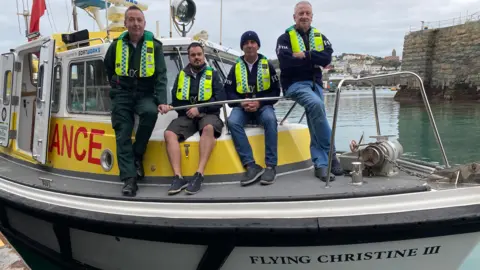 Four men wearing high-vis lifebelts are on the deck of the Flying Christine III as she is tied up alongside a harbour wall. Two of the men are sitting on the bow and two are standing. Navigational equipment and a search lights are just visible on the roof. There are houses in the distance across the sea.