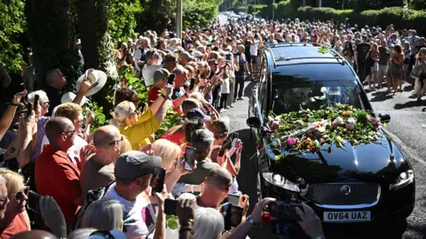 Bystanders flock towards a black hearse and throw flowers onto its bonnet as it drives through the town carrying the coffin of Bonnie Tyler.