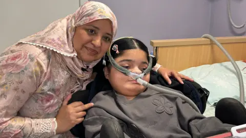 Maham looks at her iPad as she sits with her mother, Nomia. She is wearing a device which helps with ventilation which happens twice a day and through the night.