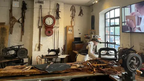 A leather work station with a man hunched over a long wooden table working on a peice of material.
