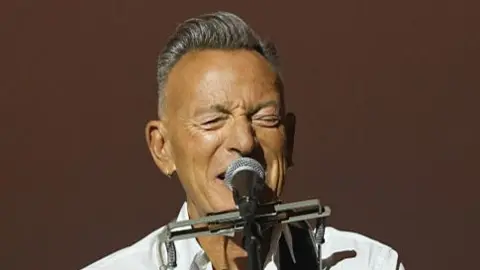 A man with short grey hair singing into a mic, wearing a white shirt.