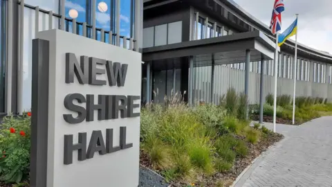 Exterior of council building with a sign saying New Shire Hall. Building is modern and there is planting of grasses and flowers around entrance. two flag- the Ukrainian and British are on poles. 