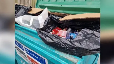 Darlington Council Black bin bags containing cigarettes and vapes sit on rubbish in a large, blue commercial wheelie bin.