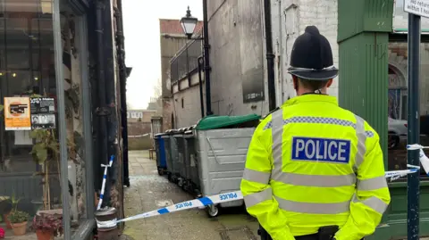 A police officer is standing looking away from the camera, on the right had side of the picture. In the middle shows an alley way with a row of 5 bins. Police cordon tape has also been placed around the scene. 