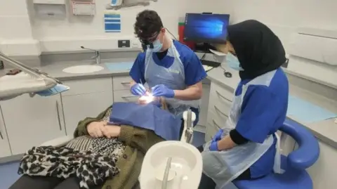 A patient is seated in a dentist's chair. A dental student wearing scrubs and PPE is looking in her mouth. Another student, also wearing scrubs and PPE, is looking on