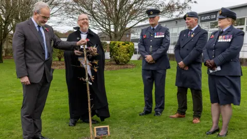 A small ceremonial gathering outdoors on the grass. Five people stand around the newly planted tree, which has a commemorative plaque at its base.