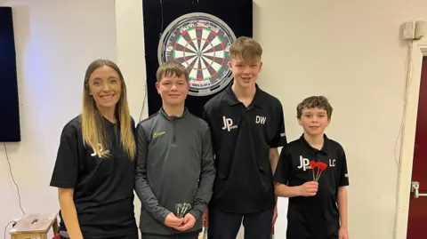 BBC Bobbi Jones next to three children who are holding darts, standing in front of a darts board