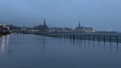 BBC Weather Watchers/Lawrence A dark view of a promenade by a beach. It is covered in water and the lights of a town can be seen in the distance