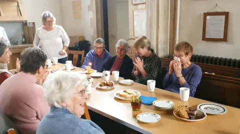 A group of older people sat around a table talking and singing happy birthday to someone