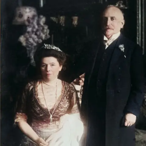 Antony Easton Colorised historical photograph of Olga and Heinrich Eisner in formal attire. Olga is seated, wearing a lace dress with a tiara and holding a white cloth. Heinrich stands beside her in a dark suit with a boutonniere. The background includes floral arrangements and ornate decor, indicating an elegant setting.