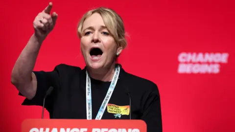 Reuters Sharon Graham - a woman with blonde hair and wearing a lanyard and black outfit - stands in front of a red wall behind a podium with a microphone, pointing in the air as she speaks.