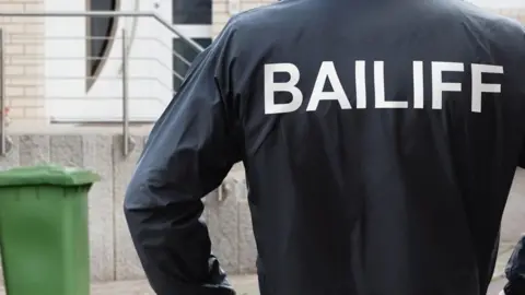 Rear view of a male bailiff standing with his hands on his hips at the entrance of a house.