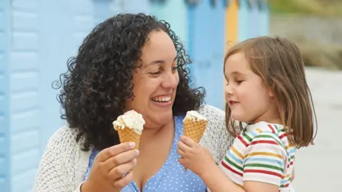 Getty Images A woman is holding a female child as they eat ice cream. The woman has black curly hair and the child has brown long hair. They are smiling in the picture. the woman is wearing a blue top with white polka dots, and a white cardigan, and the child has a white top with coloured stripes on it.