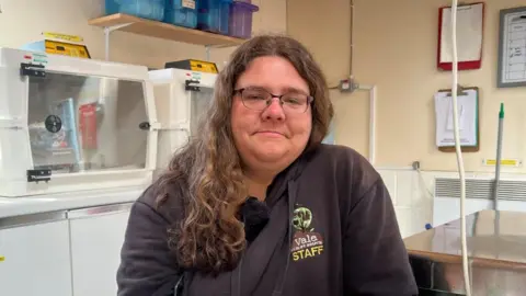 A woman with glasses and long, curly brown hair is wearing a brown polo shirt with Staff written on. She is sitting in an office surrounded by glass boxes where rescued animals are kept