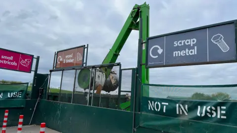 Designated zones for scrap metal, wood and timber, and small electricals at a recycling centre. There is a large, green, crane-like compactor breaking down the wood and timber. It is a cloudy day.