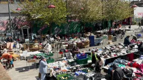 An aerial view of a market, with traders standing behind tables, talking to customers.