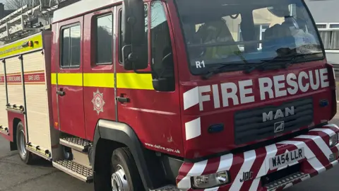The front and side of a Devon and Somerset Fire and Rescue Service fire engine parked on a hill. 
