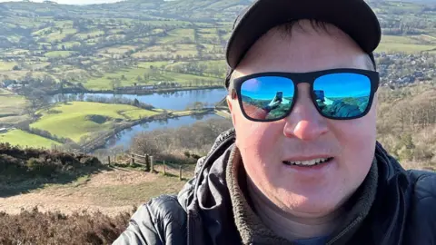 A selfie shows Ryan in clue sunglasses with black rims and a black baseball cap. he is wearing a black coat and is smiling at the camera. Behind Ryan, in the distance is field and a lake.