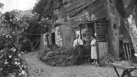 National Trust A black and white photo of a man and a woman in the doorway of a rock house, with the wooden porch leaning against a sandstone cave with cut in windows and doors. He wears a white shirt and tie and trousers with braces. She has a light dress with a pattern on it. They stand on a bricked pathway outside the house.
