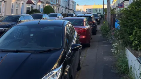 BBC A row of cars parked on a pavement on a narrow street