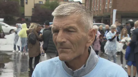 Neil McCrimmon looking slightly off camera. He has grey hair and is wearing a light blue t-shirt over a grey jumper.