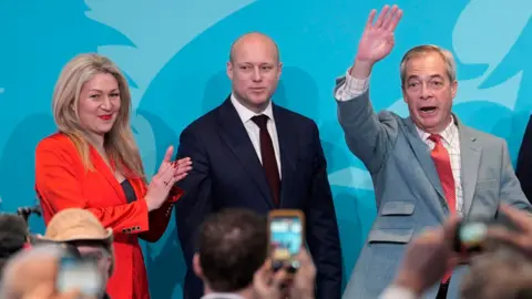 PA Media A woman with blonde hair in an orange jacket, Laura Ann Jones, a bald man in a navy suit, Dan Thomas, and a man with grey hair and a light blue suit, Nigel Farage, stand on stage at an event.