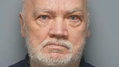 Michael Figgins glowers at the camera in a police custody photo. He has a short, white beard and his eyes are bloodshot.