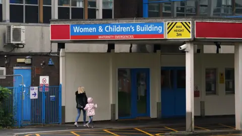 PA Media A blonde woman in a black coat and blue jeans walks into the Women & Childen's Building at the Countess of Chester Hospital holding hands with a young girl, who has a pink coat with the hood up and blue leggings. 