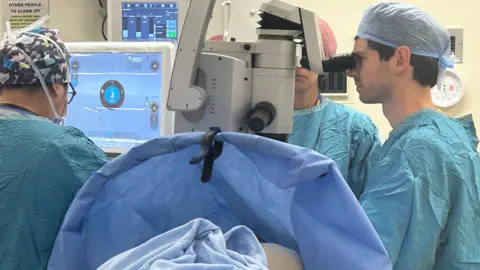 A man dressed in green scrubs looks through a microscope to perform cornea transplant surgery. The patient is lying on a hospital bed with her head underneath the microscope