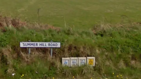 A sign at one end of the road that reads Summerhill Road. It is positioned in a grassy embankment, with a field behind it.